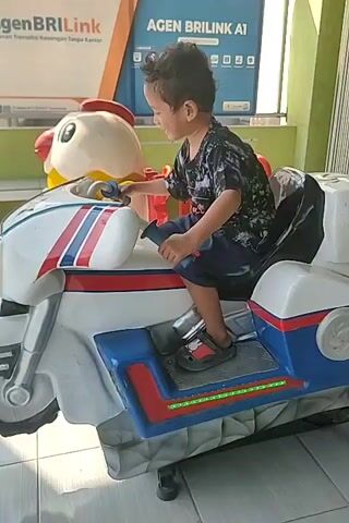 A young boy is sitting on a white toy motorcycle with blue and red accents. He is wearing a dark t-shirt with a pattern and dark shorts, and sandals. The motorcycle is positioned in front of a building with a sign that says "AGEN BRILINK A1". The boy is looking to his left and smiling. The toy motorcycle has a yellow and red chicken head on the front. The environment appears to be an outdoor area with a tiled floor and a wall in the background.