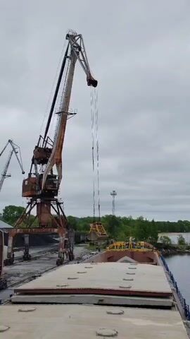 This video captures an industrial port scene in Medvezhyegorsk, Russia, under an overcast sky. The primary subject is a large, weathered port crane, exhibiting significant rust and wear, which is actively operating. Its long boom extends over the cargo area of a bulk carrier or barge, which is docked alongside.

The crane is in the process of lowering a bright yellow grab or bucket into what appears to be an open hold of the vessel, indicating ongoing loading or unloading of bulk material. The vessel has large, flat hatch covers, typical of a cargo ship. In the background, to the left, another, smaller crane is partially visible, suggesting a larger port infrastructure. Across the waterway, green trees line the distant shore, contrasting with the industrial foreground.

The sky is uniformly grey and cloudy, setting a somewhat muted tone for the scene. The numbers "4.1" are clearly visible on the base of the main crane. The machinery shows signs of age and heavy use. No people are visible in the frame, though the active operation of the crane implies human involvement. The overall scene depicts a functional, busy port environment.