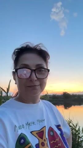 A woman with dark hair and glasses wearing a white t-shirt smiles at the camera. She is outdoors near a body of water with reeds in the foreground and a sunset in the background.