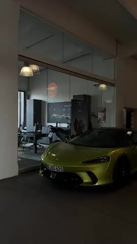 The video shows a lime green McLaren sports car parked inside a garage or showroom. The car is positioned in the foreground, with its front facing the camera. Behind the car, a large glass window reveals an interior space. Inside the visible area, there is a desk with a computer, a motorcycle, and a large screen displaying a "Gulf" logo. There are also several posters on the wall. The lighting in the scene appears to be artificial, with overhead lamps illuminating the space. To the right of the car, there are racks of clothing and another "Gulf" logo sign. A person is visible in the background behind the clothing racks. The overall impression is of a modern, well-lit automotive or retail space.
