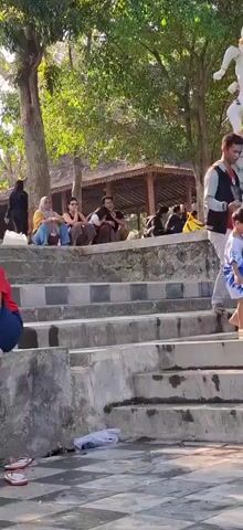 The video shows an outdoor scene with many people sitting on stone steps. Trees provide shade, and a traditional-looking building with a thatched roof is visible in the background. A statue of a figure, possibly a monkey or a deity, stands to the right. Some people are dressed in casual clothing, while others wear headscarves. A young boy in a blue and white shirt is standing on the steps. Litter is visible on the lower steps and ground. The sun is shining, casting shadows.