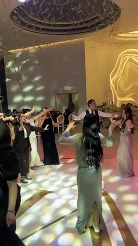 The video shows a group of people dancing in what appears to be a large hall, possibly a wedding venue or event space. The camera moves around the room, capturing groups of men and women. Some women are wearing long dresses in different colors, including black, gray, and beige. The men are mostly in white shirts or suits. The dance floor has a circular pattern with lights. The hall is decorated with a modern aesthetic, featuring tall structures with gold-colored designs. The overall atmosphere appears lively and celebratory.