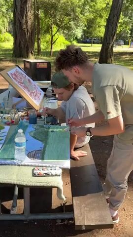Two young men are painting outdoors at a picnic table. One man, wearing a green baseball cap, is seated and actively painting on a large sheet of paper spread across the table. He is holding a paintbrush in his right hand and appears focused on his work. The other man stands behind and to the right of the seated man, observing the painting. He is also holding a paintbrush and seems to be offering guidance or assistance, with his right hand near the seated man's painting.

Their painting depicts a landscape scene with a blue sky, green grassy areas, and a building that resembles a house with a chimney. There are also small dark shapes that might represent animals or objects in the grass.

On the table are various art supplies, including tubes of paint, brushes, water containers, and palettes with different colors. Another artwork is propped up on an easel behind them, featuring abstract red and brown shapes. The setting appears to be a park or campsite, with trees and foliage visible in the background, suggesting an outdoor art session. The lighting indicates it is daytime, likely sunny.
