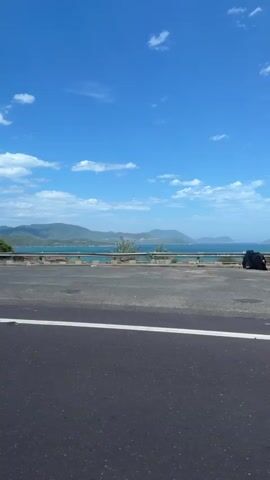 A POV shot from a moving vehicle on a road reveals a coastal landscape. The scene opens with a clear blue sky dotted with white clouds and a body of water in the distance. A road with a white line marking its edge is visible in the foreground. As the vehicle moves, the view pans slightly, showing a guardrail along the roadside. Further along, a billboard with text and images of buildings and water appears on the left. The vehicle continues, and the roadside transitions to a grassy embankment with scattered vegetation and some debris. More trees and shrubs line the landscape, with hills or mountains visible in the far distance under the bright sky. The video captures the essence of a scenic drive along a coastal route.