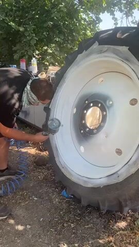 A person wearing a mask and a painter's smock is spray painting a large tractor wheel. The wheel is mostly white, with a dark gray rim around the center. The person is holding a spray gun and aiming it at the wheel. There are bottles and tools in the background, and the scene appears to be outdoors.