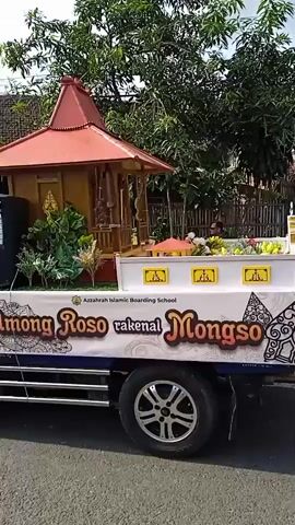 A daytime outdoor scene shows a parade moving along a street. People are sitting on the side of the road watching the parade. A truck is shown decorated with a small gazebo-like structure on the back, and the words “Rakenal Mongso” are visible on the side. People in the parade are carrying flags and signs. Many people in the parade are wearing black jackets, batik-patterned sarongs, and headbands. The scene shows shops and houses along the side of the road.  Some of the shops have signs visible. The sky is partly cloudy.