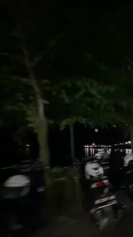 The video shows a nighttime scene of parked motorcycles along a waterside area.  The camera appears to be moving slowly past the motorcycles. Trees with lights strung between them are visible.  Reflections of lights can be seen on the water in the background.