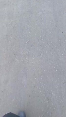 The video shows a single, dark, leather shoe walking on a gray asphalt surface. The camera appears to be held by the person wearing the shoe, as the perspective is directly down towards the ground. The surface is textured and uneven, indicating it is likely a road or a paved area. The lighting suggests it is daytime, but potentially overcast, as the shadows are soft. The shoe appears to be moving forward at a steady pace.