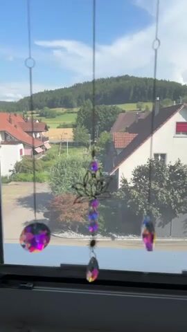 This is a vertical, medium shot video of a window with suncatchers hanging in front of it, overlooking a scenic view of a rural European village on a sunny day. The suncatchers are made of chains with colorful, faceted crystal pendants that catch the light and cast rainbows. One suncatcher in the center is more elaborate, featuring a lotus-like design with purple crystal beads.

The scene outside the window reveals a charming village with red-tiled roofs and white houses nestled amongst lush green trees and rolling hills. In the distance, a verdant forest covers the higher slopes, with patches of golden fields interspersed in the valley. The sky is a clear, bright blue with a few wispy white clouds. The sunlight suggests it is daytime, likely late morning or early afternoon.

The overall mood is peaceful and idyllic, evoking a sense of calm and natural beauty. The presence of the suncatchers adds a touch of whimsical decoration to the otherwise natural landscape. There is no visible text in the video. The setting appears to be a residential area in Lufingen, Switzerland, given the architectural style of the houses and the surrounding landscape. The focus is on the interplay of light and color from the suncatchers against the backdrop of the picturesque scenery.