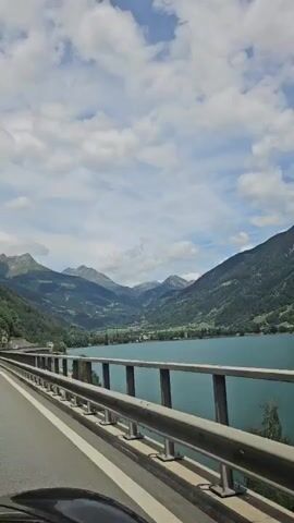 The video shows a view from a moving vehicle, likely a car, looking out over a scenic landscape. The camera is pointed forward and slightly to the right. In the foreground, there is a paved road with a white line marking the lane. A metal guardrail runs along the right side of the road, separating it from a large body of turquoise water. Beyond the water, the landscape rises into lush green mountains that are dotted with trees. The sky is partly cloudy with patches of blue visible. The sun is shining, casting light on the mountains and the water. The overall impression is one of a serene and beautiful natural environment.