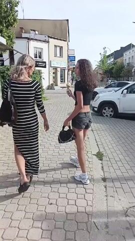 This media file captures a sunny daytime scene on a paved walkway in Piaseczno, Poland. Two individuals are the primary subjects. The first, a woman with blonde hair, walks away from the camera, wearing a long, black and white striped dress and dark sandals. She carries a dark bag over her left shoulder.

The second subject is a younger person with long, curly brown hair, also walking away from the camera. They are wearing a black crop top, denim shorts, and white sneakers. This individual is holding an ice cream cone and a black handbag. Their body language, with a slight turn of the head, suggests they might be looking back or to the side.

The setting appears to be a commercial area. Behind the subjects, there's a small kiosk or shop with a glass display window. Further down the street, a tall, blue vending machine with a prominent Pepsi logo and smaller logos for Lipton and Mirinda is visible. Several parked cars, including a yellow hatchback, are also present. Lush green trees line the background, indicating it's likely spring or summer. The overall mood seems casual and pleasant, consistent with a warm day. The presence of ice cream suggests an enjoyable outing. The time of day is clearly daytime, illuminated by bright sunlight casting shadows. No specific emotions are overtly displayed, but the activity of enjoying ice cream on a pleasant day implies contentment.
