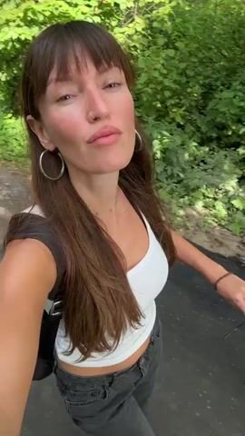 The video begins with a medium shot of a woman with long brown hair and bangs, wearing a white tank top and large hoop earrings, looking at the camera and smiling. She has a nose ring and appears to be outdoors in a wooded area with green trees. In the background, another person with blonde hair, wearing a white t-shirt and backpack, is walking away from the camera. The woman in the foreground is holding a black backpack strap and a dark object in her right hand. The camera then pans slightly to the right, showing the blonde-haired person walking further away. The video then cuts back to a close-up of the woman's face as she puts on a pair of dark sunglasses. The environment appears to be a paved path or trail surrounded by foliage.
