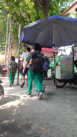 A group of children in school uniforms walk down a paved path. The children are wearing green pants and patterned shirts. Most of them are carrying backpacks. A food cart with a large blue umbrella is parked on the right side of the path. The cart has a sign that says "CIMOL". Trees and vines hang overhead, casting shadows on the path. In the background, there are motorcycles parked and a building. The sun is shining brightly.