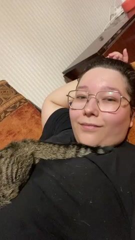 A person lies on their back, propped up by pillows on a couch. The person is wearing glasses and a black t-shirt. A tabby cat is lying on the person's chest. The cat's tail is draped over the person's shoulder. The cat is looking down at the person's shirt. The background is a wall with a patterned wallpaper.