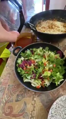 A person adds olive oil to a salad of mixed greens and chopped tomatoes in a black pan. Another pan containing cooked pasta with a creamy, cheesy sauce sits in the background. An open bottle of dark liquid sits next to the salad pan. The table on which the pans sit is decorated with a colorful, mosaic-like pattern. A yellow safety vest is also visible on the table. Sunlight streams in from an open door in the background.