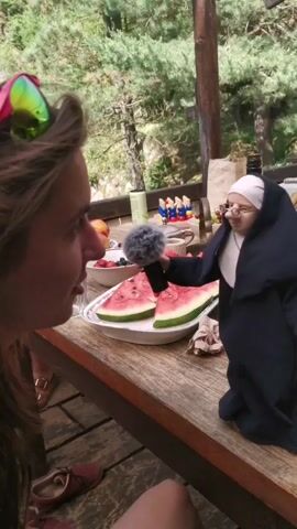 A medium shot on a sunny day shows a young woman sitting on the left with a small nun doll on the right, both looking at each other, on a wooden table. The woman, with long brown hair and colorful sunglasses on her head, is wearing a black top. The nun doll is dressed in a black habit and a white coif, holding a small black microphone with a fuzzy wind muff in its right hand. A white platter with watermelon slices is between them on the table, along with other fruits and small decorative items in the background. The scene takes place outdoors on a deck or patio, with trees and greenery visible behind them.