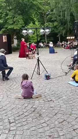 The video shows an outdoor performance taking place on a paved courtyard. In the foreground, a young girl with braided hair sits on the ground facing away from the camera, watching the performance. Behind her, a light stand with four spotlights is set up. Further back on a raised platform, three individuals are present. A woman in a long red gown stands to the left, appearing to address the other two individuals. Two men in red jackets are seated on chairs. To the right of the stage area, another woman in a blue dress and bonnet is seated. An audience is visible in the background, seated at tables and on chairs. The environment appears to be a garden or park, with trees and foliage surrounding the performance area. The overall scene suggests a theatrical or cultural event being held outdoors.