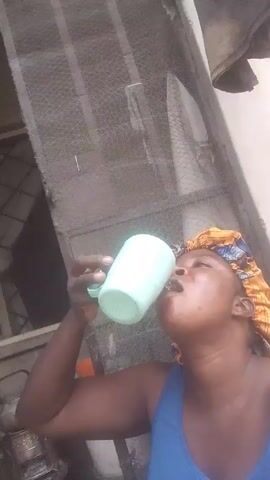 A woman is drinking from a pale green plastic mug. She is wearing a blue tank top and a yellow and blue patterned head covering. The scene is taken outdoors, with a screen door in the background. There is a metal pot on the ground next to her.