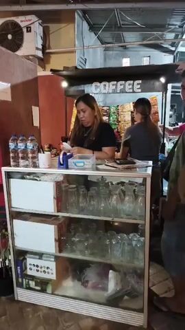The video shows a woman behind a counter with a sign that says "COFFEE." She appears to be serving a man who is standing in front of the counter. There are bottles of water and glasses on the counter. In the background, there are shelves stocked with snacks. Another woman is standing behind the counter to the left of the first woman. There are also other people visible in the background, including a person with a dog. The scene appears to be an outdoor food stall or small cafe.