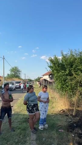 The video begins with a view of two young women standing on a grassy area next to a road. Behind them, several vehicles are parked on the road, and a group of people are walking away from the camera. The woman on the left is wearing a green t-shirt and floral shorts, while the woman on the right is wearing a white t-shirt and patterned pants. They appear to be observing something off-camera. The scene is outdoors with a clear blue sky. The video then pans to the right, showing a burnt area with blackened ground and scattered debris. A wooden fence runs along the edge of the burnt area. Further into the distance, there are trees and buildings. The camera continues to pan, revealing a larger expanse of burnt land with a thin trail of smoke rising in the distance. More people are visible standing on the grassy verge, observing the scene. The overall impression is of a rural or semi-rural area that has experienced a fire. The second half of the video shows more of the burnt landscape and people watching.