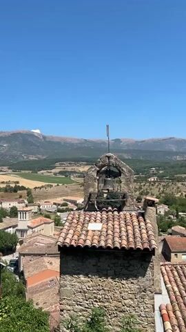 The video opens with a clear blue sky and a panoramic view of a village nestled in a valley, surrounded by rolling hills and distant mountains. In the foreground, a weathered stone bell tower with a red-tiled roof stands prominently. A flag with a black emblem on a light background flies from a pole atop the bell tower. Below the bell tower, a cluster of buildings with terracotta roofs characteristic of European villages is visible. The landscape beyond the village features agricultural fields, green trees, and more distant, rugged mountains. The lighting suggests it is a sunny day. The video captures a static scene, with no discernible movement or activity apart from the slight fluttering of the flag. The camera angle is elevated, offering a bird's-eye perspective of the village and its surroundings.