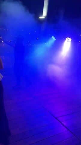 A live concert stage is set with a dense fog, illuminated by shifting white, blue, and green lights. A man in a black shirt and pants walks across the wooden platform towards the camera.  Another man walks in from the left. The music has a driving rhythm.