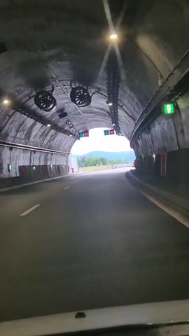 The video begins from the perspective of a vehicle inside a tunnel, showing the arched concrete ceiling with lights and ventilation fans. The road ahead is paved and curves slightly to the right. Traffic signals are visible, indicating a speed limit of 80. As the vehicle exits the tunnel, the view opens up to a highway with a cloudy sky and distant mountains. The road continues to curve, with a barrier on the right and trees and fields on the left. Other vehicles are visible in the distance.