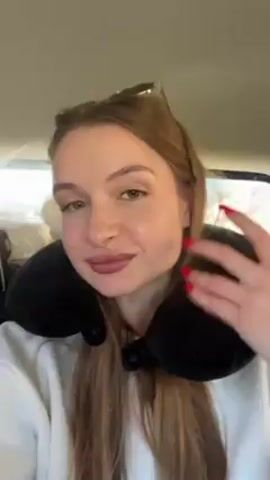 A woman with long brown hair, wearing a white sweatshirt and a black travel pillow, is sitting in a car. Her nails are painted red. She is talking and gesturing with her hands. The car appears to be moving.