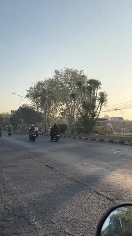 A video shows a road scene with several motorcycles and a small truck on a paved road. The motorcycles are carrying passengers, some with helmets. The truck appears to be carrying wooden crates. Trees and utility poles are visible along the side of the road. The sky is clear and appears to be early morning or late afternoon, with the sun casting long shadows. The video seems to be taken from the perspective of someone riding on a motorcycle.