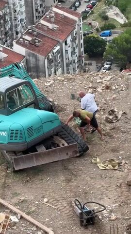 Two men are working on the track of a teal-colored excavator on a dirt slope. The man on the left, wearing a white t-shirt and blue shorts, is using a shovel to push at the track. The man on the right, wearing a green t-shirt and black shorts, is leaning over and appears to be pulling on the track with his hands. There are construction materials and debris scattered around them, including a yellow strap and what looks like a small plate compactor in the foreground. In the background, there are several apartment buildings and a street with cars.