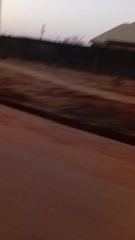 The video, taken from a moving vehicle, shows a road beside a dirt area. A tall fence lines the dirt area, with some structures visible behind it. At one point, a blurry figure of a person with a green object walks alongside the road. The sky is overcast. The primary focus is on the passing landscape next to the road.
