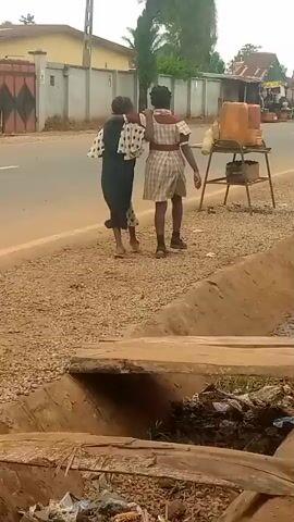 Two individuals are walking away from the camera on a dirt path alongside a paved road. The person on the left is wearing a dark dress with a white polka dot pattern on the sleeves. The person on the right is wearing a checkered shirt and shorts. They have their arms around each other's shoulders. To the right of the road, there is a metal stand holding several large, opaque orange containers. In the foreground, a drainage ditch filled with debris is visible. The background shows buildings, trees, and utility poles.