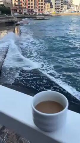 The video shows a cup of coffee on a white railing, with a view of the ocean and some buildings in the background. Waves are crashing against a stone barrier. The water is blue and choppy. The buildings are multi-story and appear to be residential or commercial. The sky is overcast. The camera is relatively stable, with slight movements.