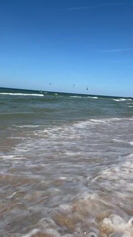 A wide shot of a beach shows gentle waves lapping the shore under a clear blue sky. In the distance, several kite surfers are visible on the water, their colorful kites dotting the horizon. The sand is a light tan color, and the water is a clear green with white foam from the waves. The sun appears to be high in the sky, casting bright light on the scene.