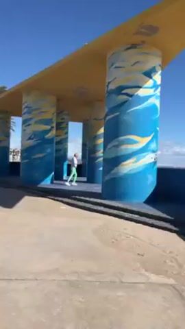 The video captures a man in a white shirt and green pants walking through a pavilion-like structure on a sunny day. The structure consists of a flat, yellow roof supported by several large, round columns, each decorated with a pattern of blue and yellow that resembles waves or clouds. The man walks back and forth across the small elevated platform inside the structure. The area outside the structure appears to be a seaside promenade, as evidenced by the concrete surface and a partial view of the sea in the background.