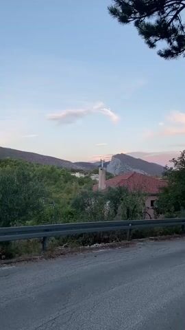The video shows a rural landscape at dusk. In the foreground, a grey asphalt road is visible, bordered by a metal guardrail. Beyond the road, lush green trees and bushes create a dense foliage. A red-tiled roof of a house peeks through the greenery, with a tall, light-colored chimney topped with what appears to be antennae or a weather vane. In the middle ground, several houses with white walls and red roofs are nestled among the trees. The background features rolling hills and a prominent, jagged grey mountain range bathed in the soft, pastel hues of the setting sun. The sky is a pale blue with wispy pink and white clouds. A faint crescent moon is visible in the upper left quadrant of the sky. The overall impression is one of tranquility and natural beauty.