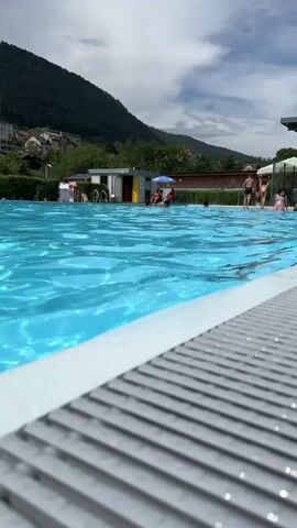 The media file shows a low-angle view of a swimming pool on a sunny day. In the foreground, a slatted pool deck leads to the bright blue water, which is rippled by movement. In the mid-ground, several people are enjoying the pool. Some are swimming, while others are relaxing on the deck or near the pool's edge. A small building with changing rooms and showers is visible, as is a blue umbrella providing shade. In the background, a lush green mountain slopes upwards, dotted with houses and trees. The sky is partly cloudy, suggesting a pleasant summer day. The scene evokes a feeling of relaxation and summer fun. The location appears to be a public swimming pool in Saint-Imier, Switzerland, given the surrounding mountainous landscape and architecture. No visible text is present in the media file.
