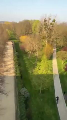 The video starts with an aerial shot of a park with a path where a person is riding a bicycle. The camera then shifts, revealing a river. Trees line the riverbank. The video seems to have been shot from a moving vehicle, as evidenced by the occasional appearance of a window frame and a small red-and-white marker at the bottom of the frame. The weather is sunny.