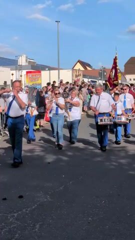 A marching band is parading down a street. Several people are playing drums and other brass instruments. The band members are wearing white shirts with blue ties and blue pants. There are spectators on the side of the street watching the parade. The sky is blue and there are some buildings in the background. One person in the parade is carrying a flag. The video captures the band from a low angle, focusing on the musicians and their instruments.