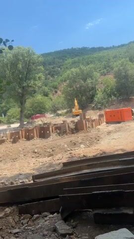 A construction site is shown with a yellow excavator in the background operating near a series of metal sheet pilings. In the middle ground, there are more sheet pilings and a bright orange generator. A large green hill covered with trees forms the background under a clear blue sky. The foreground contains debris and more metal sheeting.