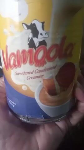 The video shows a close-up of a can of Vamoola sweetened condensed creamer. A person's hands are holding the can. The can has a yellow background with a picture of a cow, and the brand name "Vamoola" in red letters. The words "Sweetened Condensed Creamer" are written below the brand name. There is an image of a glass with a brown liquid and what appears to be chocolate in it. The person speaking describes the product as sweet and delicious.
