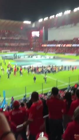 The video shows an outdoor scene at night in a packed soccer stadium.  The stadium is predominantly red, and floodlights illuminate the field.  Many people are on the green field, milling about, while spectators in red shirts and hats occupy the stands.  A large video screen is visible at one end of the stadium.  The spectators in the foreground are standing and appear to be cheering. The words "Corona" are visible on their shirts.