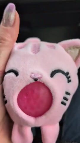 A person's hand with manicured nails holds a pink plush toy cat with black embroidered eyes and whiskers. The cat has a wide open mouth, and inside its mouth is a large, transparent pink ball filled with smaller pink beads. The hand is squeezing the toy. The background is dark and out of focus.