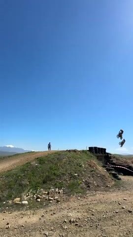 The video shows a motocross track on a sunny day with a bright blue sky. Two motorcyclists are jumping off a dirt ramp lined with black tires. One rider is in the air at the moment the video starts, and another rider is approaching the ramp behind him. A person is standing on a grassy hill overlooking the jump. The landscape in the background features dirt tracks, sparse vegetation, and distant mountains with snow on their peaks.