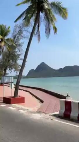 The video shows a waterfront promenade on a sunny day. The camera, possibly mounted on a moving vehicle, travels alongside a paved area with a red and white border separating it from the adjacent road. On the promenade, there are trees, including palm trees, providing shade. The turquoise water of the sea stretches out towards a mountainous island in the background under a clear blue sky. Two people can be seen sitting on a bench next to a tree, facing the water. The wind is clearly audible in the video.