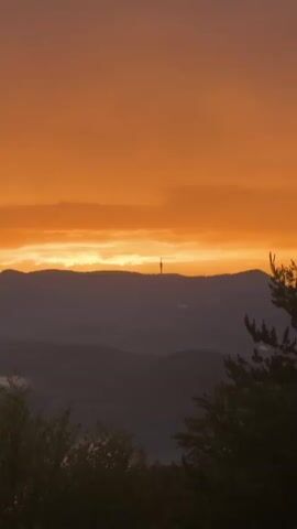 A silhouette of a radio tower stands atop a dark mountain range against a vibrant orange sunset sky. The sky is filled with streaks of clouds illuminated by the setting sun. In the foreground, the dark shapes of trees are visible. The video zooms in slightly on the radio tower throughout its duration.