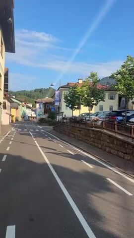 A low-angle, vertical shot pans across a sun-drenched street lined with buildings. The road is paved and has white lane markings. In the foreground, a large white arrow points left, indicating a turning lane. To the right, a bike lane is marked with a white bicycle symbol. A stone retaining wall runs along the right side of the road, topped with a metal fence. Parked cars are visible beyond the fence. The buildings on the left are multi-story structures with balconies and sloped roofs. The buildings on the right are smaller and appear to be shops or homes. Trees with green foliage are scattered throughout the scene. The sky is bright blue with a few wispy clouds, and a faint sunbeam or lens flare is visible. In the distance, hills covered in green trees rise under the sky. A silver station wagon is seen driving away from the camera on the right side of the road. The overall impression is of a quiet, picturesque European town on a clear day.