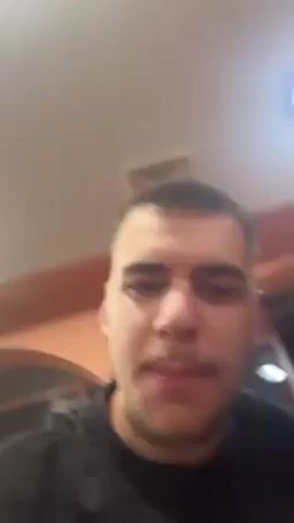A close-up video shows a young man with a buzz cut and a dark shirt singing along to music while holding up his phone to record. A young woman with long, curly, dark hair steps into the frame from the side. They both smile and pose for the camera, mouthing the lyrics and briefly doing a duck face. The background shows a corner of a room with warm lighting, an arched opening, and a window. The music appears to be playing in the background.