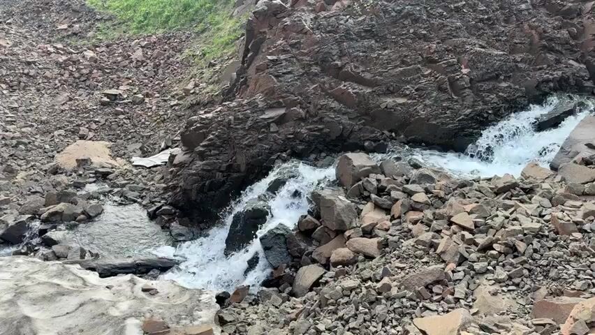 This is a video recording of a narrow, rocky gorge with a stream flowing through it. The gorge is characterized by steep, dark rock faces and a scree slope covered in loose rocks and debris. Patches of snow and ice are visible on the left side of the frame, suggesting a cold climate or recent snowfall. The stream is fast-flowing and turbulent, with white water cascading over rocks. In the background, the gorge narrows further, and sparse green vegetation can be seen clinging to the slopes. The scene appears to be in a remote, mountainous area. The time of day and weather are not clearly discernible, but the lighting suggests it could be daytime. There are no people or man-made objects visible in the video. The overall impression is one of a rugged, wild, and natural landscape. The video is shot from a static viewpoint, allowing the viewer to observe the movement of the water and the details of the rocky terrain. The recording appears to be a continuous shot without any editing or cuts.