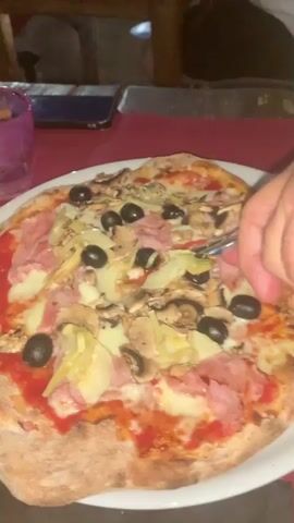A close-up shot shows a whole pizza on a white plate. The pizza has tomato sauce, cheese, ham, mushrooms, olives, and artichoke hearts. A hand is using a knife and fork to cut a slice of the pizza. The pizza is on a red tablecloth, and a purple glass is visible on the left side of the frame.